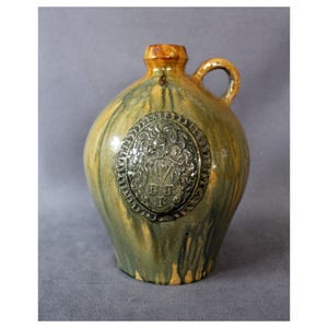 May include: A green and brown ceramic jug with a handle. The jug has a raised oval design with a crest and the letters "W.F.P." in the centre.
