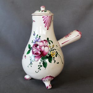 Può includere: Brocca in ceramica bianca con coperchio, manico e tre piedini. La brocca è decorata con un grande motivo floreale rosa e viola, foglie verdi e dettagli gialli. Il manico e i piedini hanno un bordo rosa.