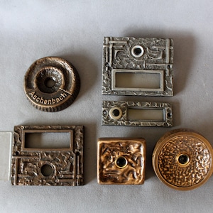 Può includere: Collezione di ferramenta per porte vintage in bronzo e metallo, tra cui maniglie, targhette e un campanello. I pezzi hanno superfici strutturate e varie forme, alcuni con il nome "Aschenbach".