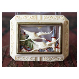 May include: Ivory-colored rectangular box with a gold-framed diorama. The diorama depicts two white birds in flight over a village with red-roofed houses and a tall church. The box has an ornate, carved border.
