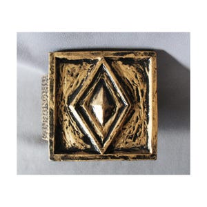 Op de afbeelding: Een vierkant, goudkleurig decoratief object met een verhoogd ruitvormig ontwerp in het midden. Het object heeft een getextureerd oppervlak en een zwarte rand. Het is waarschijnlijk een decoratief element of een kunstwerk.