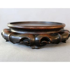 Könnte beinhalten: Ein dunkelbrauner, geschnitzter Holzständer mit einer kreisförmigen Oberseite und einem verzierten, gerollten Sockel. Der Ständer ist aus reichhaltigem, dunklem Holz gefertigt und hat eine glatte, polierte Oberfläche. Der Sockel weist eine Reihe von geschwungenen, fließenden Formen auf.