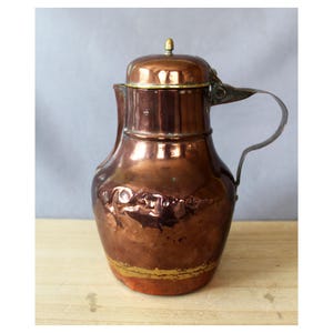 Può includere: Una brocca in rame martellato con corpo arrotondato, collo stretto e coperchio a cupola sormontato da un piccolo pomello in ottone. La brocca presenta un manico in metallo curvo fissato lateralmente e una fascia in ottone vicino alla base.