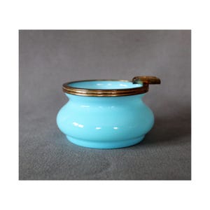 May include: A light blue, round ashtray with a gold-colored rim and a small rest. The ashtray has a bulbous shape and a smooth, glossy finish. A vintage piece for home decor.