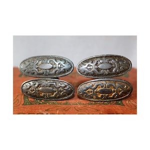 Op de afbeelding: Vier antieke ovale metalen knopen met gegraveerde patronen. De knopen hebben een zilveren afwerking met gedetailleerde ontwerpen en een vintage uitstraling. De knopen zijn in een vierkant gerangschikt. De knopen zijn waarschijnlijk van een metaallegering.