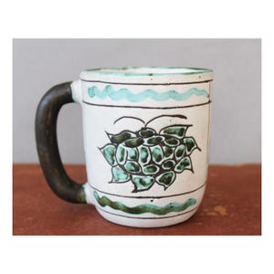 Puede incluir: Taza de cerámica blanca con asa marrón oscuro y diseño floral verde y negro. La taza presenta un patrón turquesa ondulado alrededor del borde y la base. La taza está sobre una superficie marrón.