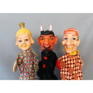 Op de afbeelding: Drie handpoppen met gevormde hoofden en stoffen lichamen. De linkerpop heeft blond haar en een bloemenjurk. De middelste pop is een rode duivel met hoorns en een zwart pak. De rechterpop draagt een oranje hemd met een geblokt patroon.