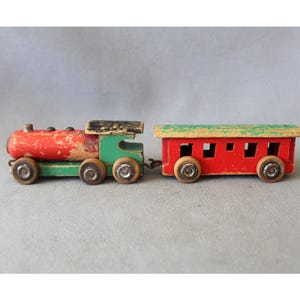 May include: A vintage wooden toy train consisting of a red and green locomotive with a black roof and a matching red passenger car with a green roof. Both pieces feature small wooden wheels and show signs of paint wear and age-related patina.