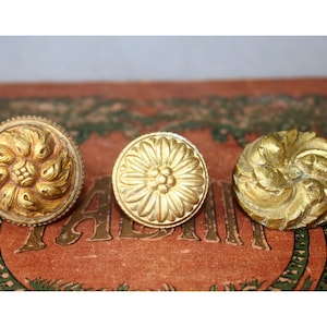 Pode incluir: Três botões metálicos circulares de tom dourado com desenhos em relevo intrincados. O botão esquerdo apresenta um padrão de folhas em espiral, o central mostra um motivo floral radial e o direito exibe uma folhagem orgânica texturizada.