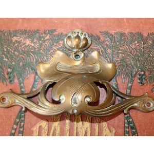 Op de afbeelding: Oud messing ladeknop met een sierlijk bloemen- en krulontwerp. De knop heeft twee montagegaten en een centraal decoratief element. De achtergrond toont een vervaagde illustratie van palmbomen en tekst.