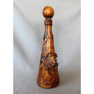 Peut inclure: Une bouteille décorative faite à la main, de forme conique et avec un bouchon rond. La bouteille est marron avec des ornements métalliques, dont une tête de lion avec un anneau. Le design présente des motifs en relief et une texture semblable au cuir.