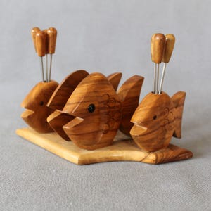 May include: A wooden fish-shaped appetizer holder with three fish figures. Each fish has an open mouth and holds small skewers. The holder is made of light brown wood and sits on a rectangular base, perfect for serving appetizers.