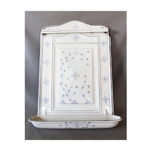 May include: A white enamelled metal wall-mounted match holder featuring a rectangular backplate with a curved top edge. The surface is decorated with delicate blue floral patterns and thin lines, and it includes a small attached tray at the base.