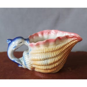 Peut inclure: Un pot à lait en céramique en forme de coquillage avec un corps texturé de couleur beige. Le bord est peint avec un dégradé rose tendre, tandis que l'anse est sculptée en forme de dauphin bleu, ajoutant une caractéristique distincte au design.
