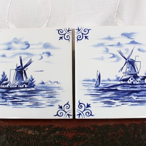 Può includere: Due piastrelle di Delft blu e bianche che raffigurano mulini a vento in un paesaggio olandese. Le piastrelle hanno uno sfondo bianco con dettagli dipinti in blu di mulini a vento, case, acqua e nuvole.