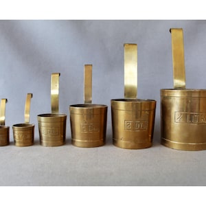 Può includere: Un set di sei misurini graduati in ottone con manici verticali. Ogni misurino presenta un testo in rilievo che ne indica la capacità, tra cui 1/2 LITRE, 2 DL, 1 DL e contrassegni più piccoli per frazioni di decilitro, disposti in fila.