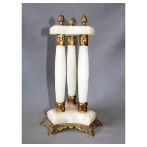 Puede incluir: Un soporte decorativo con tres columnas verticales de piedra blanca y detalles en metal dorado. La estructura incluye una base y una parte superior triangular de piedra blanca, rematadas con pies de metal dorado y tres pequeños remates.