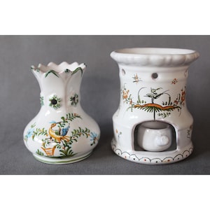 Könnte beinhalten: Zwei weiße Keramikvasen mit handbemalten Blumen- und Vogelmotiven. Die Vase links hat einen gewellten Rand und die Vase rechts hat eine runde Öffnung und eine kleine Öffnung in der Mitte.