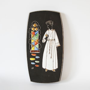 Puede incluir: Placa de pared de cerámica negra y blanca con un diseño de vitral colorido y una figura de una persona con una túnica blanca. La placa está firmada "HB".