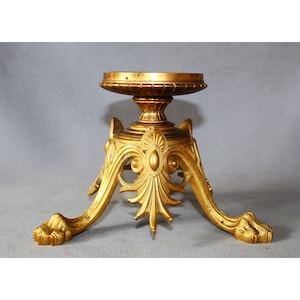 Op de afbeelding: Een goudkleurige decoratieve standaard met een ronde bovenkant en drie sierlijke poten. De poten eindigen in klauwachtige voeten. De standaard heeft een gedetailleerd, klassiek ontwerp, geschikt voor het tentoonstellen van decoratieve items.