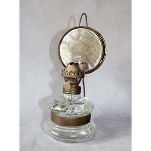Op de afbeelding: Een vintage olielamp van helder glas met een messing voet en een ronde spiegel. De lamp heeft een decoratieve messing bovenkant en een handvat om op te hangen. De spiegel is rond en gevat in een messing frame.