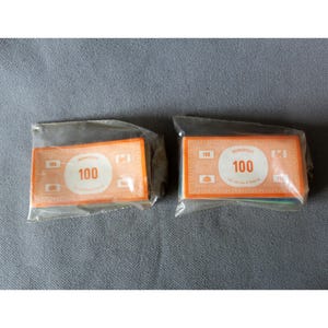 Peut inclure: Deux paquets de billets de 100 Monopoly. Chaque billet orange porte le mot "MONOPOLY" en haut, avec le chiffre "100" dans un cercle blanc. Les billets sont dans un emballage plastique transparent.