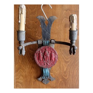 May include: A wall-mounted metal candle holder with two arms and two faux candles. The central vertical plate features a circular red medallion with embossed figures and text, accented by blue-painted details at the top and bottom of the plate.