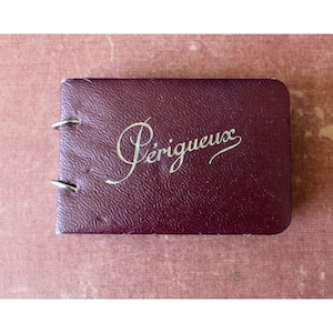 Op de afbeelding: Een klein, rechthoekig bordeauxrood boek met het woord "Périgueux" in gouden script. Het boek heeft een getextureerd oppervlak en twee zilveren ringen op de rug. De achtergrond is een gedempt roze.