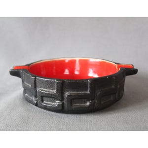 Könnte beinhalten: Ein schwarzer Aschenbecher mit geometrischem Design und leuchtend roter Innenseite. Der Aschenbecher hat zwei kleine Griffe. Das Innere ist glänzend rot und kontrastiert mit dem mattschwarzen Äußeren.