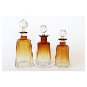 May include: Three amber-colored glass bottles with crystal stoppers. The bottles are graduated in size, with a clear base and a gradient amber hue that darkens towards the top. The stoppers are faceted and clear.