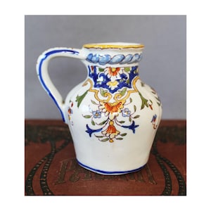 Op de afbeelding: Een kleine witte keramische kan met een blauw en geel bloemenmotief. De kan heeft een rond lichaam en een handvat.