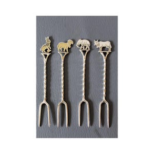 May include: Four silver-toned appetizer forks with animal-shaped toppers: a rabbit, a sheep, an elephant, and a bull. Each fork has a twisted handle and two tines. The forks are displayed on a dark gray surface.