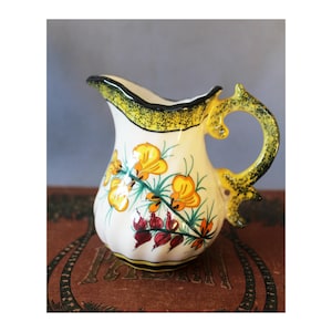 May include: A white ceramic jug featuring a hand-painted floral design with yellow and red blossoms, green stems, and a yellow rim with black speckles. The handle is yellow with black speckles and has a decorative, curved shape.