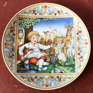 Può includere: Piatto decorativo in porcellana con una colorata illustrazione di un bambino che legge agli animali, tra cui una volpe, un cervo e una tigre. Il piatto ha un bordo dorato e un bordo con uccelli e motivi floreali. Un pezzo affascinante.