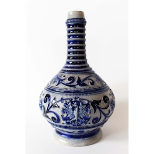 Może przedstawiać: Niebiesko-biały ceramiczny wazon o zaokrąglonym kształcie i wysokim, smukłym szyjce. Wazon jest ozdobiony misternymi wzorami kwiatowymi i herbem z ptakiem.