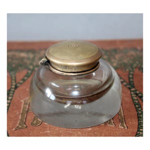 Op de afbeelding: Een inktpot van helder glas met een afgeronde, lage basis en een scharnierend, rond messing deksel. De metalen dop heeft een gladde, matte afwerking met een subtiel, gegraveerd patroon van concentrische cirkels op de bovenzijde.