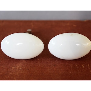 Peut inclure: Deux boutons de meuble blancs de forme ovale. Ils présentent une finition brillante et sont posés sur une surface brun-rouge foncé. Les boutons sont probablement en céramique ou en porcelaine et sont conçus pour les meubles.