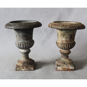 May include: A pair of weathered cast iron urns featuring a wide, fluted rim with decorative detailing, a bulbous midsection with vertical ribbing, and a square base. The metal surfaces show signs of oxidation, rust, and a mottled grey-brown finish.