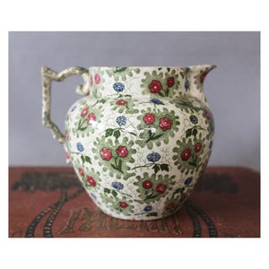 Könnte beinhalten: Eine Vintage-Keramikkrug mit floralem Muster. Der Krug hat eine cremefarbene Basis, die mit roten, grünen und blauen Blumenmustern verziert ist. Er hat einen gebogenen Griff und einen Ausguss zum Ausgießen. Der Krug wird wahrscheinlich zum Servieren von Getränken verwendet.