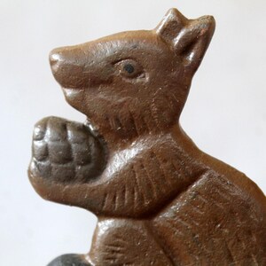 May include: A brown, cast metal squirrel figurine depicted in profile, holding a textured acorn in its front paws. The squirrel features moulded details on its body, including defined ears, a snout, and fur markings, with a smooth, dark brown finish.