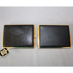 Op de afbeelding: Twee zwarte rechthoekige etuis met gouden randen. Eén heeft een textuur, reptielachtig patroon, de andere een gladde lederen uitstraling. Een klein label met Duitse tekst is aan één etui bevestigd.