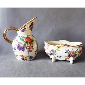 May include: A cream-coloured vintage pitcher and bowl set. The pitcher has a gold handle and rim, and the bowl has a gold rim and three small feet. Both pieces are decorated with colourful floral patterns in red, purple, blue, and yellow.