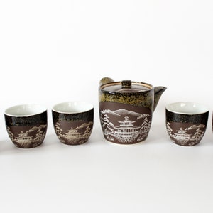 May include: A brown and green Japanese tea set with a teapot and five cups. The cups and teapot are decorated with a white design of a Japanese pagoda and trees.