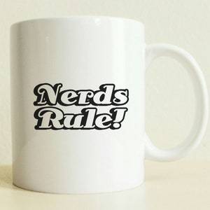Pode incluir: Caneca de cerâmica branca com texto preto que diz "Nerds Rule!" em uma fonte retrô.