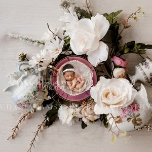 Könnte beinhalten: Ein neugeborenes Baby schläft in einer rosa Teetasse, umgeben von weißen und rosa Blumen. Die Teetasse steht auf einem goldenen Metallständer. Im Hintergrund befinden sich zwei Teekannen, eine weiße und eine blaue.