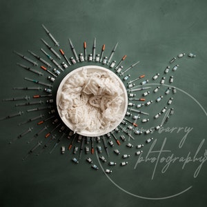 May include: A white bowl filled with white yarn, surrounded by a circle of syringes and vials on a green background. The text "emrys barry photography" is written in white on the bottom right of the image.