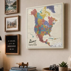 May include: A colorful map of North America hangs on a wall, surrounded by framed art and decor. The map is a detailed representation of the continent, with each state or province distinguished by a different color. A sign reads "Oh Darling, Let's Be Adventurers."