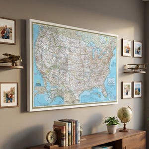 May include: A large framed map of the United States hangs on a wall, with a white border and a detailed depiction of the country's geography. The room also features framed photos, a model airplane, a globe, and a wooden cabinet.