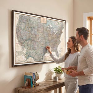 May include: A framed vintage map of the United States hangs on a wall. A couple points at the map, which is framed in dark wood. A wooden table holds books, a globe, and a mug. The map has a small plaque that reads "The Family."