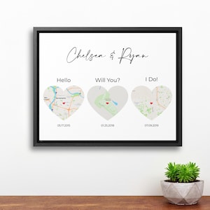 May include: A framed print with a white background and black frame. The print features the names "Chelsea & Ryan" in cursive, with three heart-shaped map illustrations labeled "Hello," "Will You?," and "I Do!" with dates below.